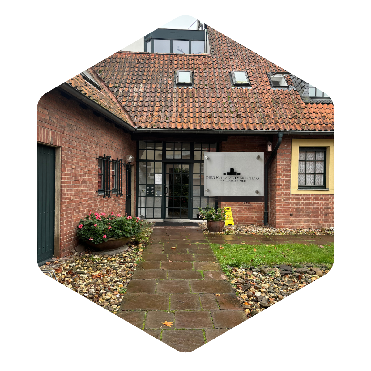Eingang der DeutscheStadtmarketing GmbH Büro in einem historischen Backsteingebäude mit rotem Ziegeldach, Glasfront und gepflegtem Außenbereich.
