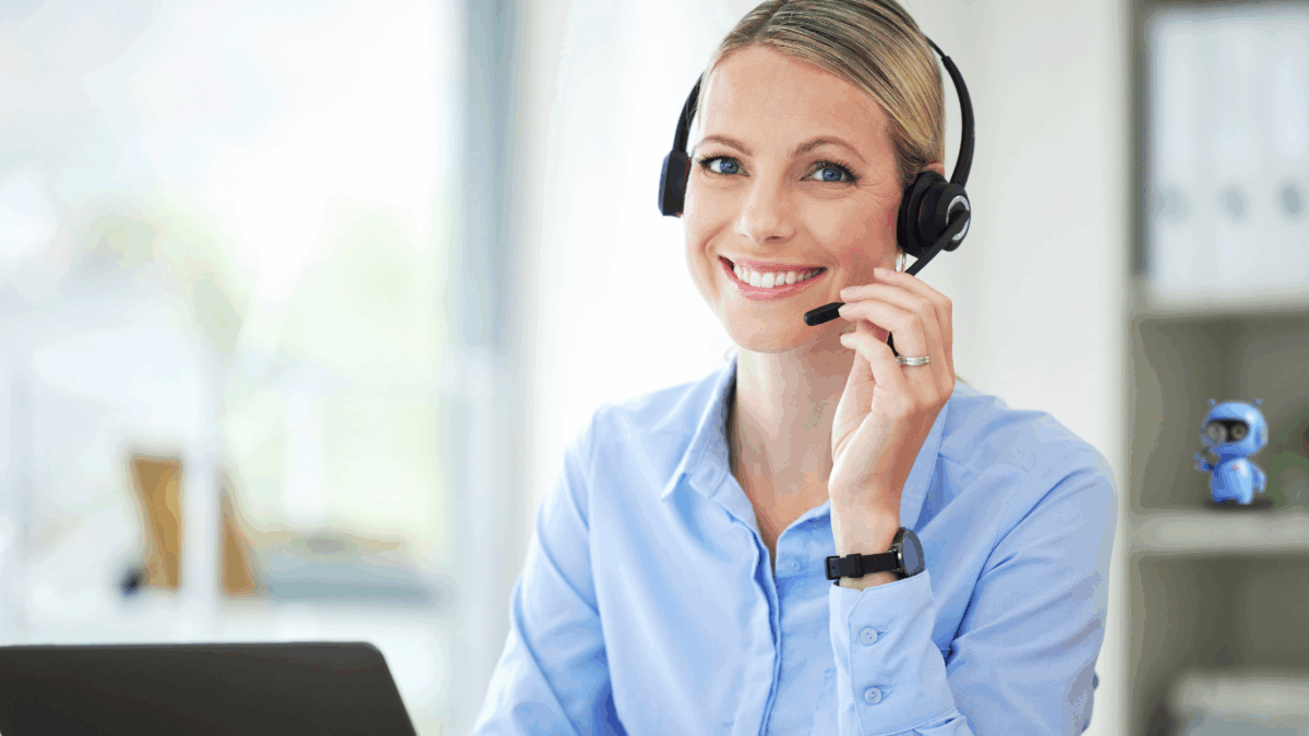 Freundliche Frau mit Headset sitzt am Schreibtisch, lächelt und spricht über ein Mikrofon.