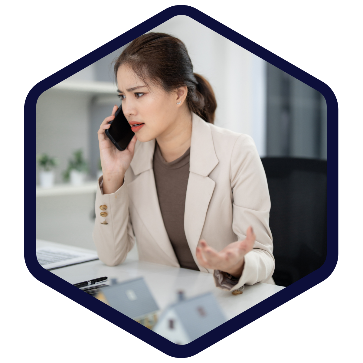 Frau in beigefarbenem Blazer telefoniert am Schreibtisch und wirkt besorgt oder gestikulierend; auf dem Tisch stehen kleine Modellhäuser.